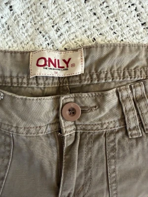 Beiga/bruna cargobyxor från ONLY - Snygga beiga cargobyxor från ONLY med raka ben, flera fickor och knappdetaljer bak. Tillverkade i ett mjukt bomullsmaterial som är skönt att bära. Perfekta för en avslappnad och trendig look.