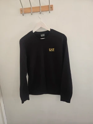 Svart EA7 sweatshirt med guld logga - Svart långärmad sweatshirt från EA7 med guld logotyp på bröstet och gula ränder i nacken. Mycket gott skick, prövad nångång