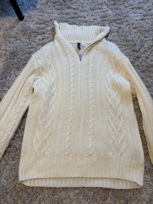 Vit kabelstickad tröja med halvzip - Kabelstickad vit tröja med halv dragkedja och hög krage. Tröjan har ett klassiskt flätmönster framtill och ribbade muddar vid ärmslut och nederkant. Perfekt för lager på lager och en clean look.