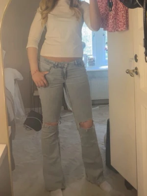 Ljusblå bootcut jeans med slitningar-Nelly - Säljer ett par ljusblå bootcut jeans med slitna knän och broderade bakfickor med röd/vit zigzag-mönster. Jeansen har låg midja och klassisk femficksdesign och är från Nelly. Strl M/38