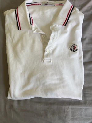 Vit Moncler pikéskjorta med ränder - Vit pikéskjorta från Moncler med klassisk krage och knappslå framtill. Kragen och ärmsluten har snygga röda och blå ränder. Broderad Moncler-logga på bröstet ger en exklusiv touch. Tillverkad i mjuk bomull för skön känsla.