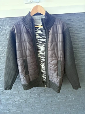 Svart stickad cardigan med quiltade detaljer - Svart cardiganjacka med stickade ärmar och rygg samt quiltad framsida. Jackan har hög krage, dragkedja och två fickor framtill. Insidan har ett zebramönstrat foder som ger en cool kontrast. Perfekt för lager-på-lager-stil. Priser kan ändras vid snabb o smidig affär.