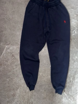 Svarta mjukisbyxor från Polo Ralph Lauren - Svarta mjukisbyxor från Polo Ralph Lauren med röd broderad logga på ena benet. Byxorna har resår och dragsko i midjan, samt muddar vid bensluten. Tillverkade i mjukt och bekvämt material, perfekta för en avslappnad stil.