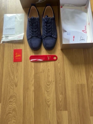 Mörkblå sneakers från Louboutin - Snygga mörkblå sneakers från Louboutin i mocka med klassisk röd sula. Skorna har låga skaft, rund tå och matchande blå skosnören. Kommer med originalkartong, dustbag och tillbehör. Perfekta för dig som vill ha något exklusivt och stilrent.