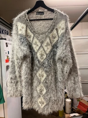 Grå fluffig stickad tröja med mönster - Supermjuk och fluffig grå stickad tröja med V-format mönster av vita rutor och små stjärndetaljer framtill. Tröjan har lång ärm och en lös, avslappnad passform. Perfekt för dig som gillar retrostil och vill ha något riktigt mysigt.