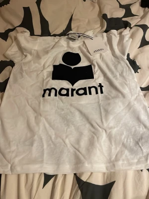 Isabel Marant T-shirt - Snygg vit t-shirt från Marant med stort svart logotryck på bröstet. Klassisk passform och rund halsringning. Perfekt för en trendig och avslappnad stil. 