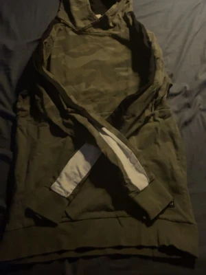 Olivgrön hoodie med camomönster - Snygg olivgrön hoodie med diskret camomönster på bröst och rygg. Hoodien har huva och ribbade muddar vid ärmslut och nederkant. Ärmarna har en vit detalj på insidan som ger en cool kontrast. Perfekt för en avslappnad streetstyle-look.