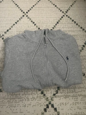 Grå zip hoodie med huva - Snygg grå hoodie med dragkedja och huva. Tröjan har dragsko i huvan, två fickor framtill och en liten mörkblå logga broderad på bröstet. Tillverkad i mjukt sweatshirtmaterial som är perfekt för chill dagar.