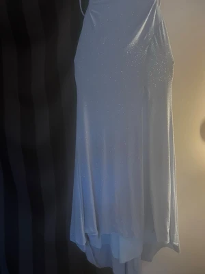 Glittrig ljusblå lång balklänning - Säljer en glittrig ljusblå långklänning med tunna axelband och asymmetrisk nederkant. Klänningen har en elegant siluett och knytning i ryggen. Perfekt för dig som vill sticka ut med en stilren och festlig look. Storlek 38. Hör av er vid frågor!