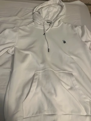 Vit hoodie med broderad logga - Snygg vit hoodie med huva och dragsnören. På bröstet finns en liten broderad logga i mörk färg som ger en clean look. Hoodien har en stor magficka och ribbade muddar vid ärmslut och nederkant. Perfekt för en avslappnad och stilren outfit. Helt äkta. Aldrig använt så den är även helt ny. Skriv för fler bilder! Priset kan diskuteras.