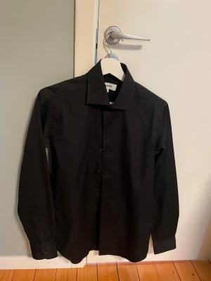 Svart klassisk skjorta med knappar - Stilren svart skjorta med lång ärm och klassisk krage. Skjortan har knappar framtill och är tillverkad i ett slätt tyg som ger en clean look. Perfekt för dig som gillar minimalistisk stil och vill ha en enkel skjorta i garderoben.