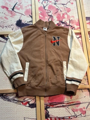 Nike Varsity Bomber Jacket - Nike Varsity Bomber Jacket i brun retro collegiate-design. Använd men i mycket bra skick. Inga synliga tecken på användning. Storlek M.  Klassisk varsity jacket med premiumkänsla, perfekt för streetwear, vintage, Y2K, college, retro och casual outfits. Snygg bomberjacka för höst, vår och vardagsbruk. Modell: DZ2551-256. Nypris ca 1250 kr.  Nike Varsity Bomber Jacket in brown retro collegiate style. Used but in very good condition. No visible signs of wear. Size M.  Classic varsity jacket with a 