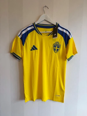 Svensk landslagströja Adidas gul/blå - Gul och blå fotbollströja från Adidas med Sveriges landslagsemblem på bröstet. Tröjan har korta ärmar, blå detaljer och klassiska Adidas-ränder på axlarna. Perfekt för match eller träning. Tillverkad i lätt och ventilerande material.