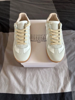 Maison Margiela vita sneakers - Stilrena sneakers från Maison Margiela i vitt och beige med klassisk låg siluett. Ovandelen är i en mix av läder och mocka, med beige snörning och gummisula. Perfekta för dig som gillar minimalistisk och clean stil.