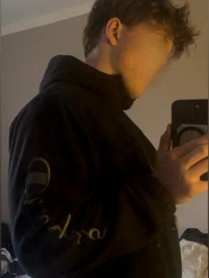 Svart Champion hoodie med tryck - Säljer en svart hoodie från Champion med text på bröstet och ärmen. Hoodien har en dragkedja vid halsen samt en ficka på bröstet. Klassisk huva och relaxed fit, perfekt för dig som gillar streetwear. Hoodien har en enkel men snygg design med diskreta detaljer. 