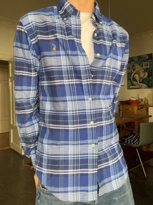 Blårutig skjorta från Polo Ralph Lauren - Säljer en snygg blårutig skjorta från Polo Ralph Lauren med button-down krage och klassisk passform. Skjortan har vita knappar och den ikoniska broderade loggan på bröstet. Tillverkad i mjuk bomull och perfekt för en avslappnad men stilren look.