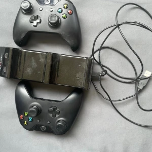 Svarta Xbox-handkontroller med laddstation - Två svarta Xbox-handkontroller med klassisk design och färgade knappar. Medföljer gör en svart laddstation och USB-kabel för enkel laddning. Perfekt för gaming med kompisar eller långa spelsessioner. Handkontrollerna har en ergonomisk form och är tillverkade i plast.