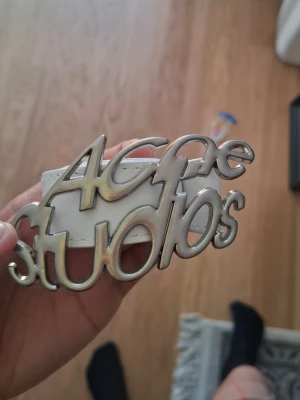 Vitt Acne Studios bälte i skinn - Säljer ett vitt bälte från Acne Studios med en stor, silverfärgad buckle i form av loggan. Bältet är gjort i skinn och har en clean, modern look som sticker ut. Perfekt accessoar för att lyfta din outfit.