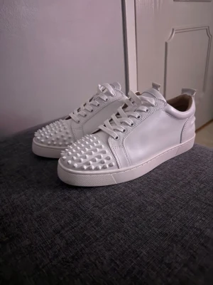 Vita sneakers med nitar från Louboutin - Säljer ett par vita sneakers från Christian Louboutin med coola nitar på tån och klassisk röd sula. Skorna är i läder och har en clean, modern look med vita snören och låg profil. Perfekta för dig som vill sticka ut med din stil. Säljer då jag inte riktigt gillade de