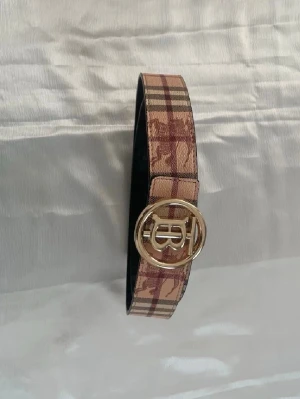 Rutigt bälte med guldspänne - Snyggt bälte med klassiskt rutigt mönster i beige, brunt och rött. Bältet har ett stilrent guldspänne med rund form och dekorativ detalj framtill. Perfekt accessoar för att lyfta din outfit.