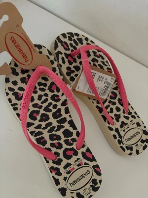 Havaianas sandaler leopardmönster rosa - Snygga flip-flops från Havaianas med leopardmönstrad sula i beige, svart och rosa samt rosa remmar med logga. Tillverkade i slitstark gummi, perfekta för sommaren eller stranden. Ikonisk design som sticker ut och ger en cool vibe till din outfit.