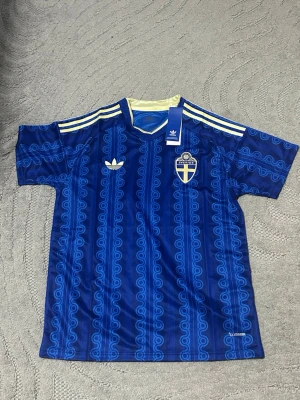 Sverige 2026 Vm bortamatch tröja storlek L  - Säljer en blå fotbollströja från Adidas med Sveriges landslagsemblem på bröstet. Tröjan har ett mönster i olika blå nyanser, gula detaljer vid krage och axlar samt Adidas klassiska ränder. Materialet är lätt och andas, perfekt för träning eller match.