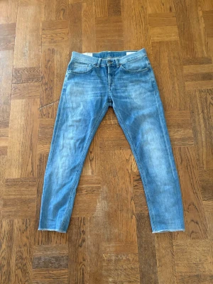 Blå smala jeans med slitningar - Säljer ett par ljusblå jeans med smal passform och snygga slitningar. Jeansen har klassiska fem fickor, orangea sömmar och en broderad detalj på bakfickan. Perfekta för en avslappnad och trendig look.