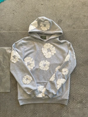 Grå hoodie med vita blommor Denim Tears - Säljer en grå hoodie från Denim Tears med vita blommönster tryckta över hela tröjan. Hoodien har huva och ribbade muddar vid ärmslut och nederkant. Perfekt för dig som gillar streetwear och vill sticka ut lite extra.