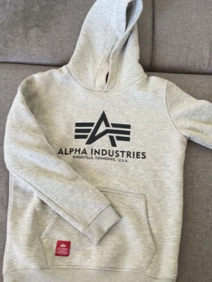  Hoodie Alpha Industries - Snygg ljusgrå hoodie från Alpha Industries med stor logga och text på bröstet. Klassisk känguruficka framtill och huva med röd detalj på insidan. Tillverkad i mjukt sweatshirtmaterial, perfekt för en avslappnad stil. Har en liten fläck men syns ej så mycket.