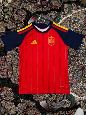 Spaniens matchtröja Adidas röd - Säljer en matchtröja för Spaniens landslag från Adidas. Tröjan är röd med gula detaljer och mörkblå ärmar, har broderad landslagssköld och Adidas-logga på bröstet. Tillverkad i lätt och ventilerande material, perfekt för fotbollsträning eller match.