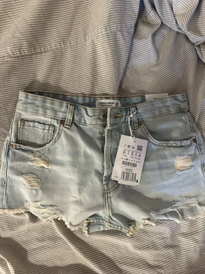 Ljusa denimshorts från Pull&Bear - Säljer ett par ljusblå denimshorts från Pull&Bear med slitna detaljer och fransig kant. Klassisk femficksmodell med bälteshällor och knappgylf. Perfekta för sommaren och ger en avslappnad vibe. Aldrig använda!! 