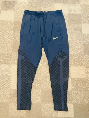 Blå träningsbyxor från Nike - Snygga blå träningsbyxor från Nike med elastisk midja och dragsko. Byxorna har ett gult Nike-märke på låret och coola mönstrade partier på benen. Tillverkade i ett lätt och stretchigt material som passar perfekt för träning eller löpning.