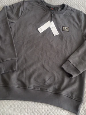 Grå sweatshirt från Calvin Klein - Snygg grå sweatshirt från Calvin Klein med rund halsringning och ribbade muddar vid ärmslut och nederkant. Tröjan har en diskret CK-logga på bröstet och är tillverkad i ekologisk bomull för extra komfort. Perfekt för en avslappnad och stilren look.