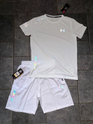 Vitt träningsset från Under Armour - Säljer ett vitt träningsset från Under Armour med t-shirt och shorts. Setet har reflekterande loggor och detaljer i regnbågsfärger. Tillverkat i ett lätt och snabbtorkande material som passar perfekt för träning och sportaktiviteter.