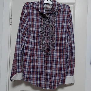 Rutig skjorta med volang från The Shirt Factory - Säljer en rutig skjorta designad av Linnéa Braun för The Shirt Factory. Skjortan har röda, blå och vita rutor, långa ärmar och snygga volangdetaljer framtill. Klassisk krage och vita manschetter ger en extra touch. Perfekt för dig som vill sticka ut lite extra.