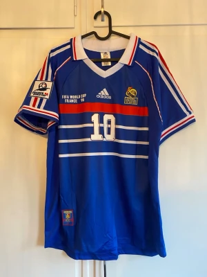 Frankrike Zidane VM 98 matchtröja - Klassisk blå fotbollströja från Adidas, Frankrikes landslag, VM 1998. Nummer 10 och Zidane på ryggen, vita och röda detaljer, officiella FIFA World Cup och FFF-emblem. V-ringad krage och korta ärmar med ränder. Perfekt för samlare eller fans.