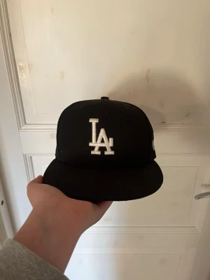 Svart LA-keps från New Era 59FIFTY - Säljer en svart keps från New Era 59FIFTY med LA-logga broderad i vitt framtill. Klassisk flat brim och stängd baksida. Märkt med Genuine Merchandise och storlek 7 1/8 (56.8 cm). Perfekt för dig som gillar streetwear och sportig stil. Hör av dig vid frågor🙌🏼