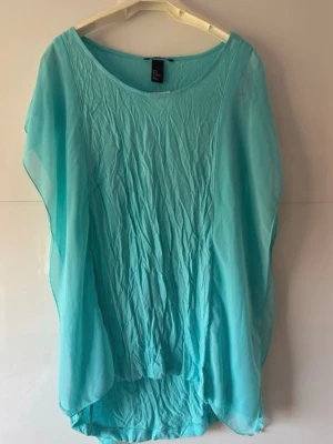  Ny.Turkos blus i chiffong - Säljer en luftig turkos blus i transparent chiffong. Tunikan har vid passform, rund halsringning och faller snyggt över kroppen. Perfekt att slänga över bikinin på stranden. Modellen är som fjäril.