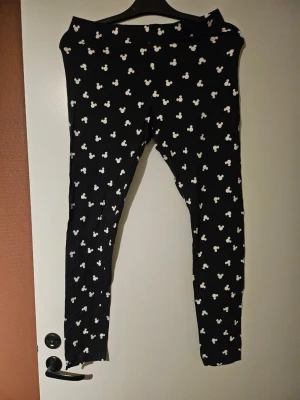 Svarta leggings med Musse Pigg-mönster - Säljer ett par svarta pyjamas leggings med vita Musse Pigg-huvuden över hela tyget. De har en klassisk, tight passform och resår i midjan. Perfekta för dig som gillar Disney och vill ha något bekvämt och lekfullt i garderoben.