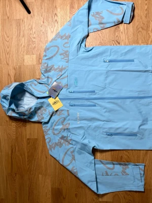 Cortiez ljus blå vind jacka  - Snygg ljusblå vindjacka från cortiez med grått grafiskt mönster på huva och ärmar. Jackan har flera dragkedjeförsedda fickor framtill, justerbar huva och kardborre vid ärmslut. Perfekt för blåsiga dagar och streetwear-stil. Passar M åsso 