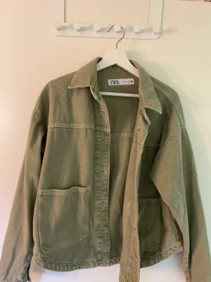 Jeansjacka från Zara - Grön jeansjacka från Zara med avslappnad passform. Jackan har två stora fickor framtill, klassisk krage och knäppning med metallknappar. Perfekt för dig som gillar en enkel men trendig look.