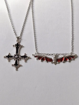 Två halsband med goth och fladdermus - Säljer två halsband i silverfärgat rostfritt stål.Det ena har ett gotiskt kors med svart emalj och stjärndetalj i mitten. Det andra har ett hänge som föreställer en fladdermus med röd emalj och fina detaljer. Perfekt för dig som gillar alternativ stil. 