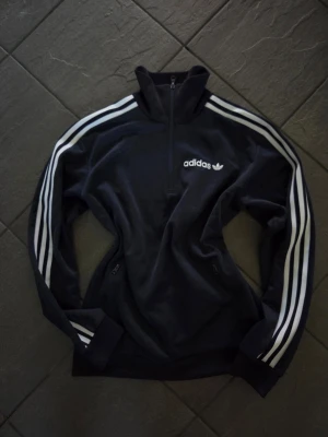 Mörkblå Adidas zip med vita ränder - Mörkblå zip hoodie från Adidas med klassiska vita ränder längs ärmarna och Adidas-logga på bröstet. Tröjan har dragkedja framtill och två fickor med dragkedja. Perfekt för en sportig och avslappnad stil.
