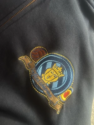 Mörkblå hoodie med broderad patch - Säljer en mörkblå hoodie med en stor, detaljerad broderad patch på bröstet. Patchen har ett blått och gult motiv med krona, boll och textband. Hoodien har synliga sömmar i kontrastfärg och är gjord i mjukt bomullsmaterial.