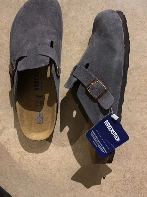Grå mockasandaler från Birkenstock - Säljer ett par gråa sandaler från Birkenstock i mjuk mocka med justerbart spänne i metall. Klassisk slip-in modell med bekväm fotbädd och robust sula. Perfekta för dig som gillar stilren och avslappnad look.