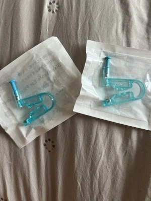 Engångs öronhåltagningsverktyg blå - Säljer två engångs öronhåltagningsverktyg i genomskinlig blå plast. De är förpackade i sterila förpackningar och har en kompakt, böjd form med metallspets. Perfekt för dig som vill ta hål i öronen hemma på ett enkelt sätt.