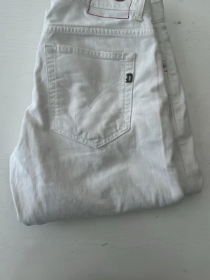 Vita dondup jeans skinny/slim fit - Säljer ett par vita jeans med rak passform och klassisk femficksdesign. Jeansen har en enkel bakficka med diskret detalj och är tillverkade i ett mjukt denimtyg. Perfekta för en clean och fräsch look.