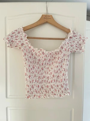 Blommig smockad topp från Bubbleroom - Supersöt smockad topp från Bubbleroom i vitt med små rosa blommor. Toppen har korta ärmar, volangkant och en söt, feminin look. Perfekt för dig som gillar romantisk stil och vill ha något luftigt och bekvämt.