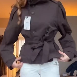 Kort brun trenchcoat med knytskärp - Snygg kort brun trenchcoat med knytskärp i midjan och axelklaffar. Jackan har dubbelknäppning framtill och klassiska detaljer som ger en stilren look. Perfekt för dig som vill ha en trendig och tidlös jacka till din garderob.