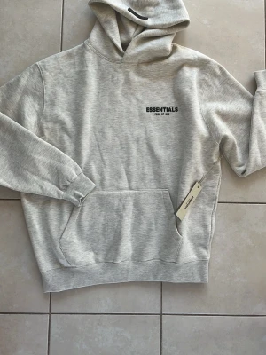 Grå Essentials Fear of God hoodie - En helt ny ljusgrå hoodie från Essentials Fear of God med svart logga på bröstet. Klassisk känguruficka framtill och huva med dragsko. Tillverkad i mjukt material som känns skönt mot huden. Perfekt för en avslappnad och trendig look. Kom gärna och skriv vid frågor och skicka gärna bud! Hoodien är i M fast är lite oversized och som passar L också! 
